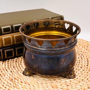 May include: A round, dark-toned metal bowl with a polished interior and a decorative rim featuring a series of cut-out heart shapes. The bowl is supported by three ornate, claw-style feet and exhibits a weathered, aged patina on its exterior surface.
