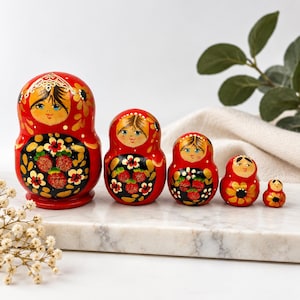 May include: A set of five red nesting dolls arranged by size from largest to smallest. Each doll features a hand-painted face with blue eyes, a black apron section decorated with red strawberries and gold floral accents, and a gold headpiece.