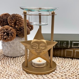 May include: A gold-coloured metal essential oil diffuser with a star-shaped face design. The diffuser has a glass dish and a small candle holder. A lit candle is inside the holder. The diffuser is on a woven mat.