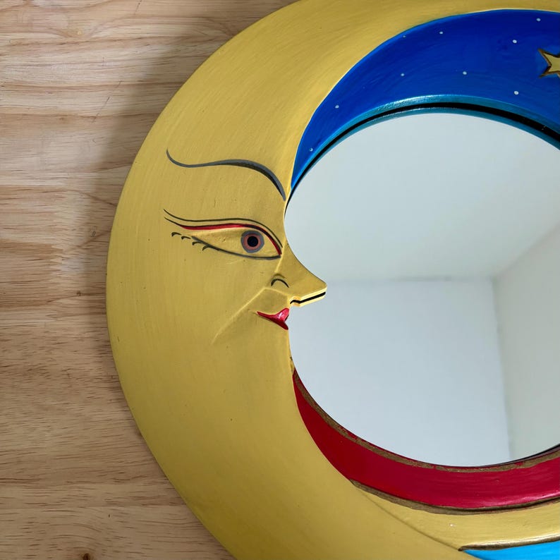 May include: A decorative moon-shaped mirror with a yellow face, blue and red accents, and a starry night sky design. The mirror has a smiling face with a red lip and eye details. The mirror is mounted on a wooden surface.
