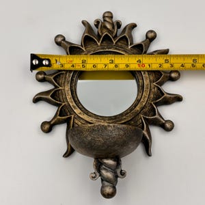 May include: A bronze-colored sunburst wall mirror with a circular center and a semi-circular shelf at the base. A yellow measuring tape is positioned across the mirror, showing a width of approximately 6 inches at the widest point.