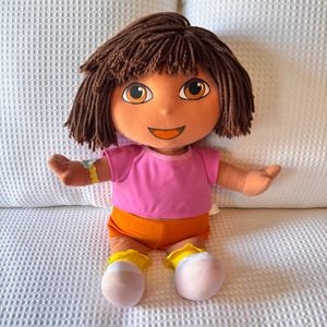 Könnte beinhalten: Plüschpuppe einer Zeichentrickfigur mit braunen Haaren, orangefarbener Haut und großen braunen Augen. Die Puppe trägt ein rosa T-Shirt, orangefarbene Shorts und gelb-weiße Schuhe. Die Puppe sitzt mit ausgestreckten Armen.