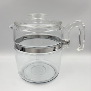May include: A clear glass percolator coffee pot featuring a matching glass lid, a sturdy glass handle, and a polished metal band encircling the upper body. The glass body displays the embossed number 9 and the brand name Stovetop clearly visible.
