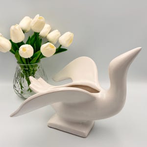 Peut inclure: Un cache-pot en céramique couleur crème en forme de cygne aux ailes déployées et à la finition lisse. À côté du cygne se trouve un vase en verre transparent texturé rempli d'un bouquet de tulipes artificielles blanches aux tiges vertes.