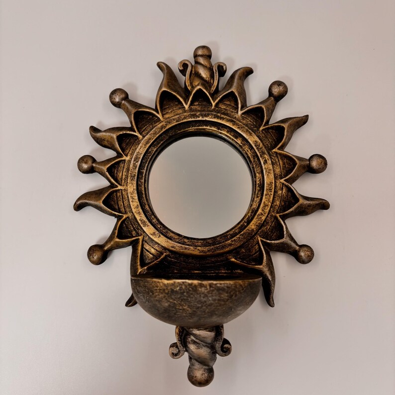 May include: A wall-mounted mirror featuring a circular glass center surrounded by a sunburst frame with pointed rays and spherical tips. The frame has a distressed bronze finish and includes a small, semi-circular bowl at the base.