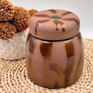 Pode incluir: Um pote de cerâmica artesanal com tampa. O pote é de cor castanha quente com riscas verticais castanhas mais escuras e um acabamento brilhante. A tampa apresenta um desenho floral e gotas de esmalte. O pote está sobre uma esteira tecida.