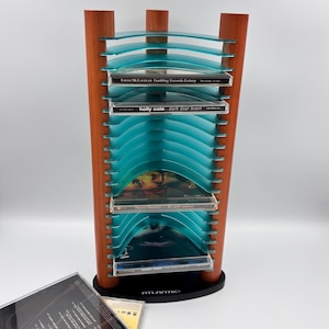 May include: A vertical media storage rack with three brown wooden pillars and multiple curved, translucent teal plastic shelves. The rack holds several CD cases, including Sarah McLachlan and Holly Cole albums, and features the brand name Atlantic.