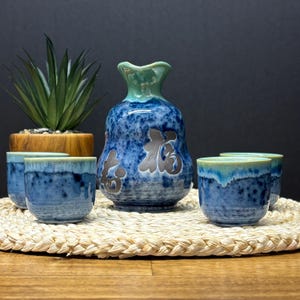 May include: A ceramic sake set featuring a blue and white speckled bottle with Japanese characters and four matching cups. The bottle has a green top, and the cups have a blue and green rim. The set is displayed on a woven mat.
