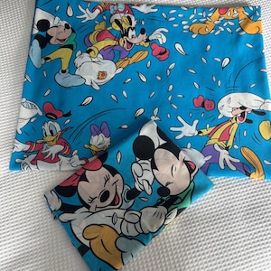 Könnte beinhalten: Zwei blaue Kissenbezüge mit einem Cartoon-Aufdruck mit Micky Maus, Minnie Maus, Donald Duck, Daisy Duck und Goofy. Die Charaktere sind in verschiedenen Posen vor einem Hintergrund aus weißen Formen. Die Kissenbezüge liegen auf einer weißen Oberfläche.