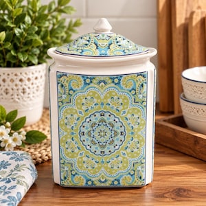 Peut inclure: Un pot en céramique avec couvercle amovible, de forme carrée aux coins arrondis. L'extérieur est orné d'un motif détaillé inspiré des mandalas en bleu, vert citron et blanc sur fond blanc, avec une finition émaillée brillante.