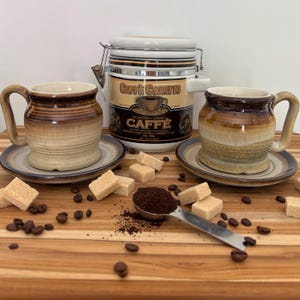 Peut inclure: Nature morte sur le thème du café avec deux tasses en céramique et leurs soucoupes, une boîte à café, des morceaux de sucre, des grains de café et une cuillère de café moulu. Les tasses et la boîte sont beiges et marron.