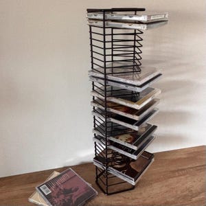 May include: A vertical black wire rack designed for storing compact discs, featuring multiple horizontal slots. Several CD cases are stacked within the rack, and one additional CD case lies flat on the surface in the foreground.