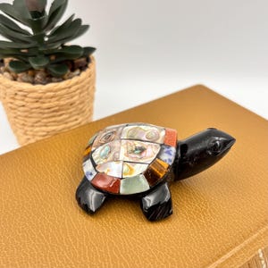 May include: A turtle figurine with a smooth black stone head and legs. The shell is crafted from a mosaic of various polished gemstones and iridescent abalone shell pieces, featuring shades of red, brown, white, and green in a geometric pattern.