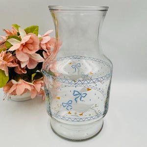 Peut inclure: Un vase en verre transparent orné d'une bande décorative avec un motif géométrique bleu, des nœuds bleu clair et des oies blanches au bec orange. Le vase présente une base large, un col effilé et un bord évasé, près de fleurs roses.