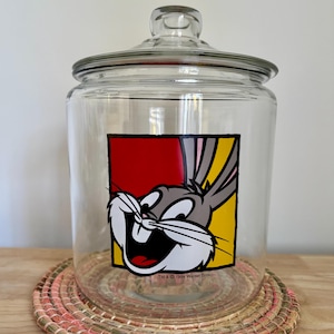 Peut inclure: Un bocal en verre transparent avec un couvercle, orné d'un dessin de Bugs Bunny sur le devant. Le dessin est placé sur un fond rouge et jaune. Le bocal est posé sur un dessous de verre tressé. Le texte "TM & © 1994 Warner Bros." est visible.