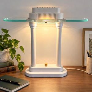 Könnte beinhalten: Eine weiße Tischleuchte mit zwei vertikalen zylindrischen Stützen, einem rechteckigen Sockel und einer horizontalen Glasablage, die aus der Mitte herausragt. Die Lampe strahlt warmes Licht unter dem oberen Teil mit Metallgitter aus.