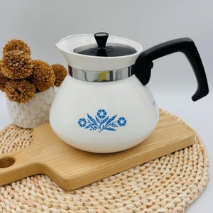 May include: A white teapot with a black lid and handle, featuring a blue floral pattern on the front. The teapot has a metallic silver band around the top section and a rounded body shape, presented as a kitchen item for brewing and serving tea.