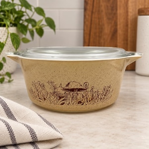 Op de afbeelding: Een ronde, beige, gespikkelde ovenschaal met een doorzichtig glazen deksel. De buitenkant is voorzien van een bruine, gedetailleerde lijntekening van diverse paddenstoelen en grassen rondom de basis. De schaal heeft twee kleine, geïntegreerde handvatten.