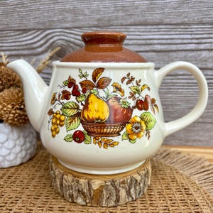 Pode incluir: Bule de cerâmica creme com tampa e alça marrons. O bule é decorado com uma cesta de frutas, incluindo peras, maçãs e uvas. O bule está sobre um descanso de madeira.