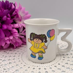 Peut inclure: Mug en céramique blanche avec une fillette de dessin animé en robe jaune tenant des ballons. La tasse a une poignée distinctive et une illustration vintage. Une fleur violette est en arrière-plan.