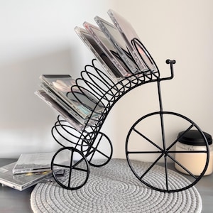 May include: Black metal bicycle-shaped CD holder. The holder has a curved back with multiple slots to hold CDs. The bicycle has three wheels and a decorative handlebar. Several CDs are displayed in the holder.