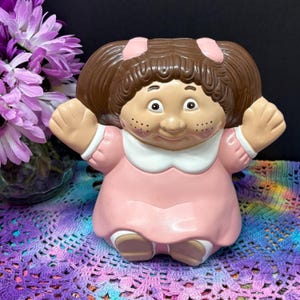 Puede incluir: Una maceta de cerámica vintage con forma de muñeca infantil, con cabello castaño, lazos rosas, un vestido rosa con cuello blanco y pecas. La muñeca tiene los brazos extendidos y está diseñada para sostener plantas.