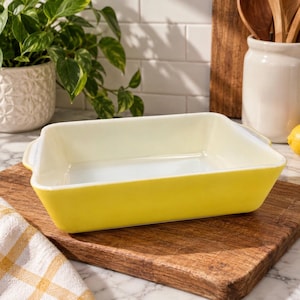 May include: A rectangular baking dish with a bright yellow exterior and a smooth white interior. The dish features integrated handles on two sides and has a deep, open basin design suitable for oven use, showcasing a clean and simple aesthetic.