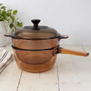 Pode incluir: Uma panela de vidro translúcido na cor âmbar com tampa a condizer. O utensílio de cozinha apresenta um cabo longo de um lado e duas pequenas pegas laterais no corpo da panela. O vidro é liso e tingido, permitindo visibilidade através dele.