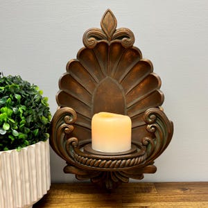 Könnte beinhalten: Ein dekorativer Wandleuchter mit einer Kerze. Der Leuchter besteht aus bronzefarbenem Metall und hat ein fächerförmiges Design mit verschnörkelten Verzierungen. Eine cremefarbene Kerze befindet sich in der Mitte. Der Leuchter ist an einer grauen Wand befestigt.