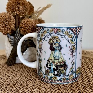 May include: White ceramic mug with a whimsical illustration of a mother and children, surrounded by sunflowers and the text "Mothers Love Grows Forever." The mug has a curved handle and a decorative border.