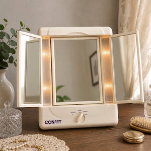 以下が含まれることがあります： クリーム色のConair製三面鏡で、垂直の照明パネルが組み込まれています。ベース部分には中央にコントロールダイヤルがあり、Off、Evening、Home、Office、Dayの各設定が記載されています。鏡には調整可能なサイドパネルが2つ付いています。