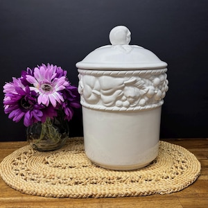 Puede incluir: Un bote de cerámica blanco con tapa, con una banda decorativa de frutas y hojas esculpidas. La tapa está rematada con un pequeño pomo redondo. Un ramo de flores moradas está a la izquierda.