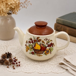 May include: A cream-colored ceramic teapot with a brown lid, featuring a colorful illustration of a basket filled with pears, apples, and grapes surrounded by green leaves and small yellow flowers on the front of the rounded body.