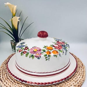 May include: A white ceramic cake dome and matching plate featuring hand-painted floral motifs in shades of pink, orange, blue, and green. The lid has a rounded red knob, and both the plate and lid edges are decorated with a scalloped red trim.