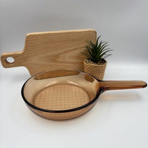 Pode incluir: Uma frigideira redonda de vidro cor de âmbar com um cabo longo, apresentando um padrão de grade texturizado na base interna. A frigideira está posicionada em frente a uma tábua de corte de madeira clara e uma pequena planta verde num vaso trançado.