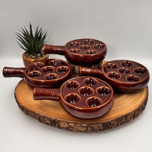 Peut inclure: Quatre plats à escargots en céramique émaillée brun foncé, chacun doté de six alvéoles circulaires et d'une courte poignée. Les plats sont disposés en groupe, présentant une finition brillante et une couleur brun profond uniforme.