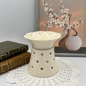 May include: Cream-colored ceramic oil warmer with a decorative, openwork design. The top dish has a scalloped edge with heart-shaped cutouts. The base features tulip-shaped cutouts. The warmer sits on a white lace doily, with books and a floral painting in the background.