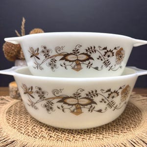 Puede incluir: Dos fuentes de horno blancas vintage con estampados florales marrones. Cada fuente tiene un asa a cada lado. El diseño floral incluye flores y hojas estilizadas en tonos marrones y dorados. Las fuentes están apiladas una encima de la otra.