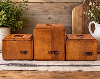 Könnte beinhalten: Ein Set aus drei quadratischen Vorratsbehältern aus Holz mit Deckeln. Der linke Behälter ist mit "TEA", der mittlere höhere Behälter mit "FARINE FLOUR" und der rechte Behälter mit "CAFE COFFEE" beschriftet. Jeder Deckel hat einen runden Griffausschnitt.