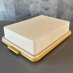 Op de afbeelding: Een rechthoekige, crèmekleurige plastic voedselopslagcontainer met een gele rand en een bijpassende gele sluiting. Het deksel heeft een licht afgeronde bovenkant en een kleine cirkelvormige inkeping. De container is ontworpen voor voedselopslag.