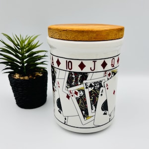Peut inclure: Un pot en céramique blanc de forme cylindrique avec un couvercle en bois naturel. L'extérieur présente un motif répétitif de diverses cartes à jouer, incluant des rois, des dames et des as, avec des enseignes rouges et noires imprimées.