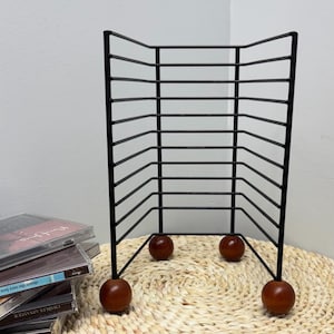 May include: A black metal CD rack with a geometric design. The rack has a series of horizontal bars and is supported by four wooden spheres. The rack is designed to hold multiple CDs. The rack is sitting on a woven mat.