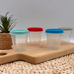 Peut inclure: Quatre petits récipients en plastique transparent avec des couvercles bleu clair, rouge, bleu et gris. Chaque récipient présente des graduations imprimées, avec le troisième gobelet affichant clairement les lignes de 45 ml et 30 ml.