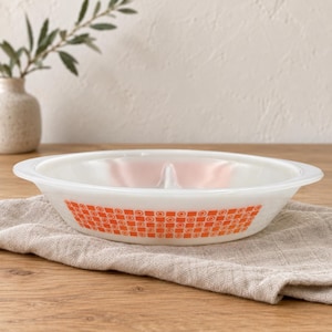 Op de afbeelding: Een verdeelde serveerschaal van wit melkglas met een herhalend oranje geometrisch patroon van vierkanten en cirkels rond de buitenrand. De ondiepe kom heeft een centrale verdeler en brede, platte handvatten aan weerszijden van de schaal.