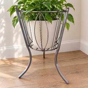 Peut inclure: Un support pour plantes en métal argenté avec une base tripode et une structure en fil de fer ornée d'accents verticaux ondulés. Le support contient un pot en céramique cylindrique blanc cassé avec une plante pothos aux feuilles vertes.