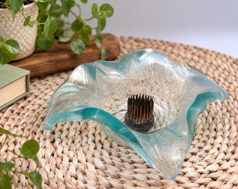 May include: A clear, wavy-edged glass bowl featuring a textured, frosted interior surface. Centered inside the bowl is a circular, dark metal flower frog with numerous upright, thin, needle-like spikes designed for arranging floral stems.