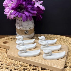 May include: Six white ceramic duck-shaped chopstick rests arranged on a wooden cutting board. The ducks are smooth and glossy, with a simple, elegant design. A vase of purple flowers sits in the background.