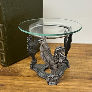 Könnte beinhalten: Ein dekorativer Kerzenhalter aus Metall mit einem Hahn-Motiv und einer durchsichtigen Glasschale. Der Kerzenhalter hat zwei detaillierte Hahnenfiguren, die eine kreisförmige Glasplatte tragen. Das Metall hat eine dunkle, antike Oberfläche, und das Glas ist transparent.