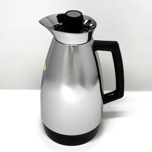 May include: A vintage chrome-plated thermal carafe with a black handle and lid. The carafe has a sleek, polished finish and a black base. The spout is designed for easy pouring. A classic piece for serving hot beverages.