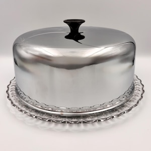 May include: A round cake dome with a polished chrome finish and a black knob handle, resting on a clear glass plate featuring a scalloped edge design. The reflective metal cover sits securely over the transparent glass base for food display.
